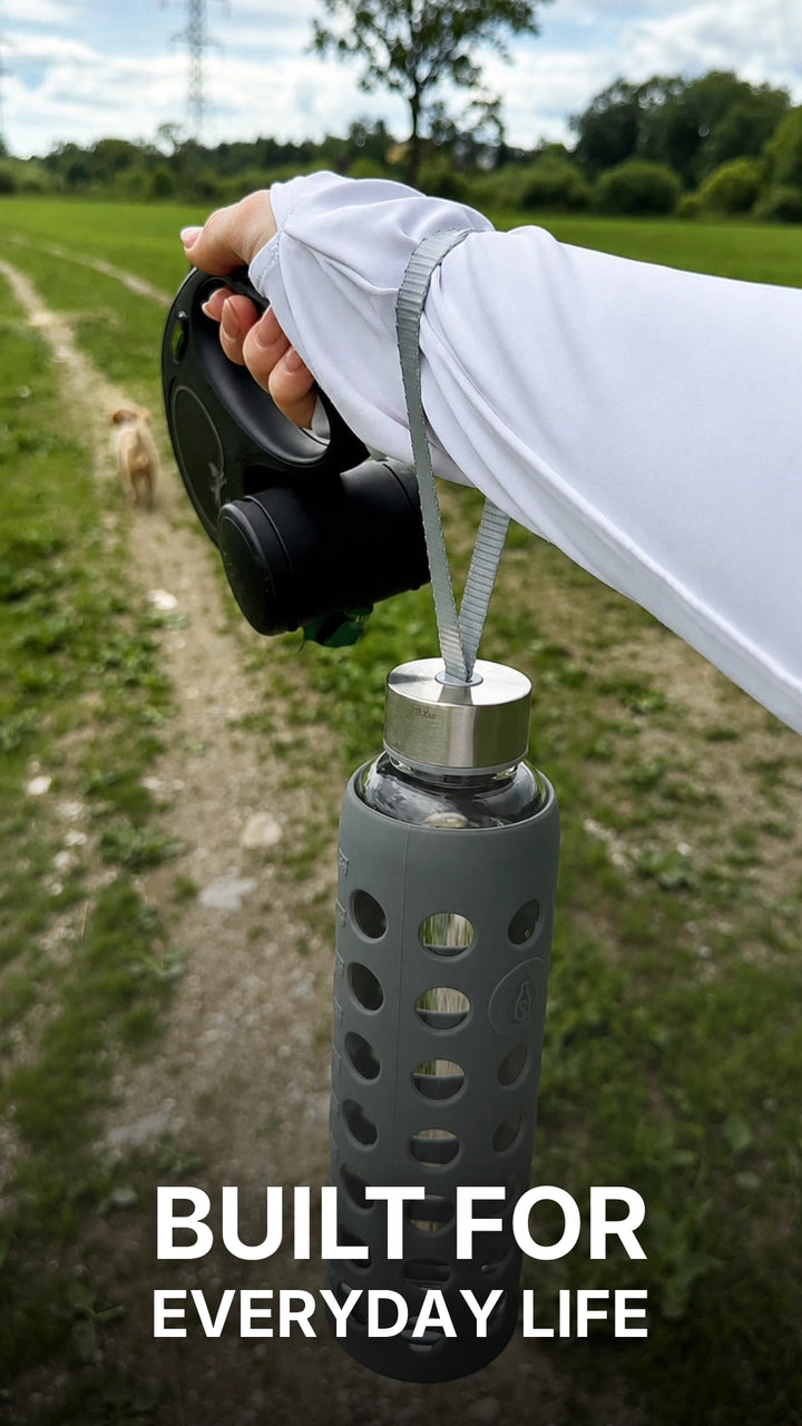 A woman stands in a grassy field and shares her experience with the spottle glass water bottle featuring a silicone sleeve. Close-up shots highlight the bottle, its wide drinking opening, and the practical carrying strap. She explains that the silicone sleeve provides a comfortable grip while adding extra protection for the glass bottle during everyday use. The video ends with a close-up of the bottle as she smiles and speaks positively to the camera.